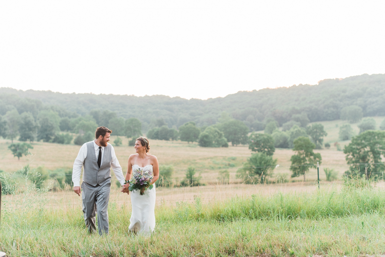 Bride and groom walking in field at Little Piney Lodge in Hermann, MO. Bride in strapless sweetheart lace wedding gown with purple bouquet with succulents. Groom in gray suit with black tie.