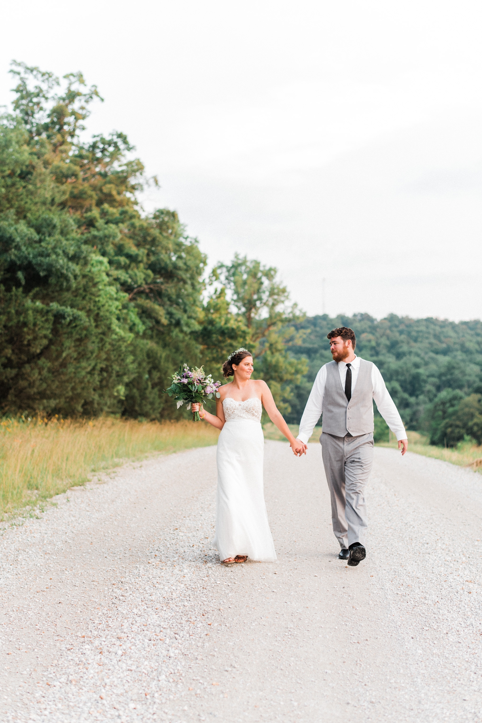 Bride and groom walking along road at Little Piney Lodge in Hermann, MO. Bride in strapless sweetheart lace wedding gown groom in gray suit with black tie.