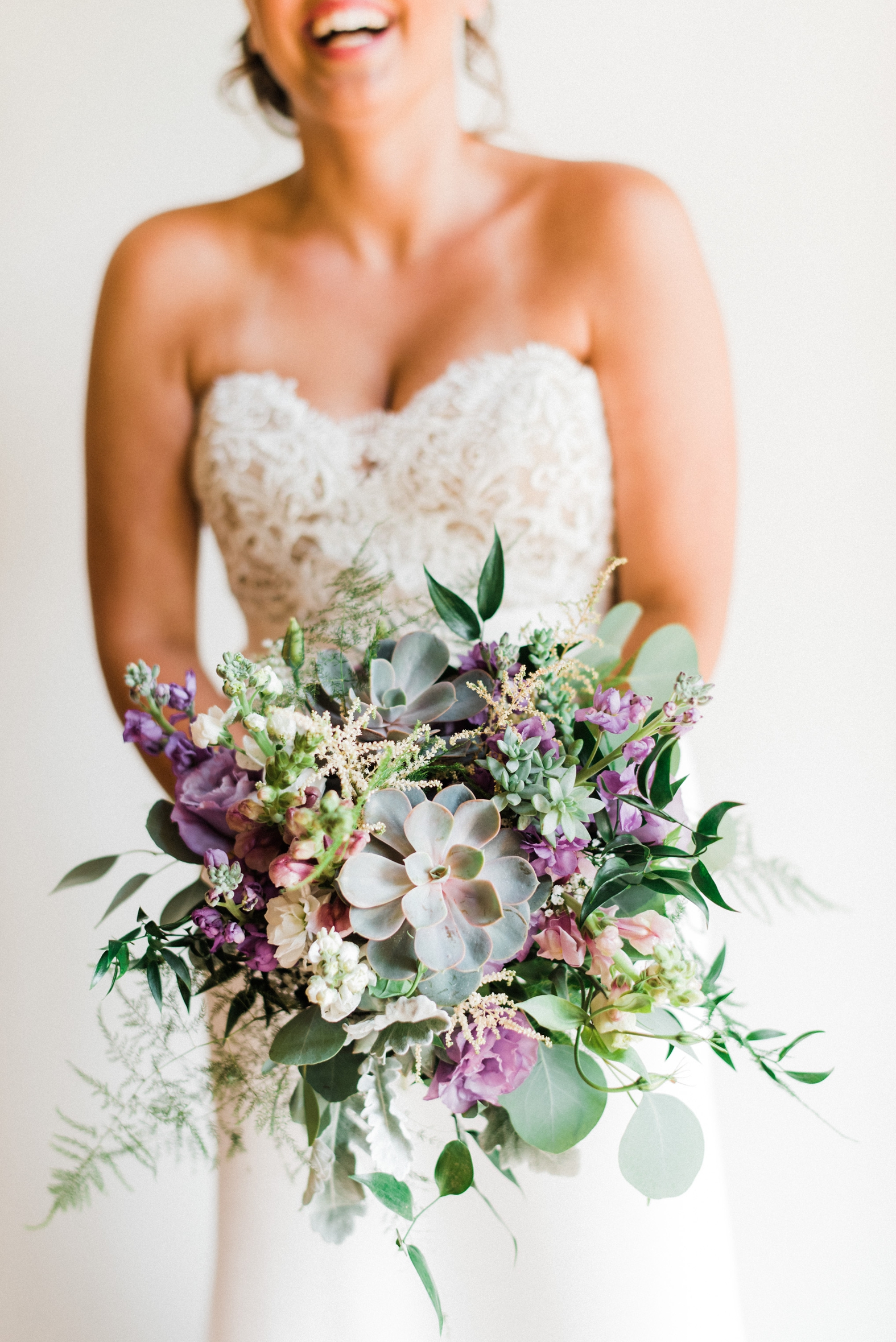 Bride laughing in sweetheart strapless lace wedding gown holding purple, lavender, and greenery bridal bouquet with succulents