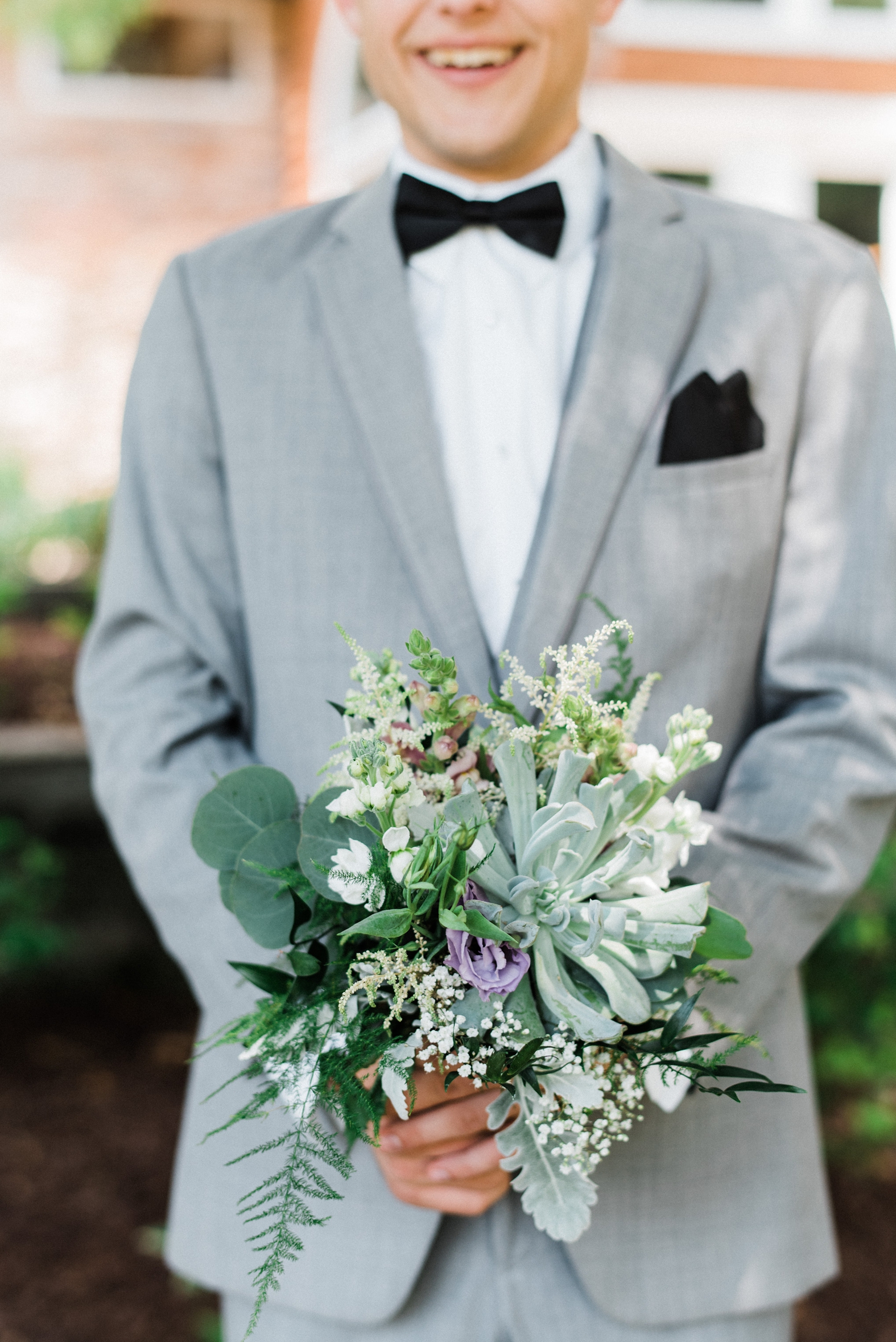 groomsman in gray suit holding bridesmaids purple, lavender, and succulent bouquet