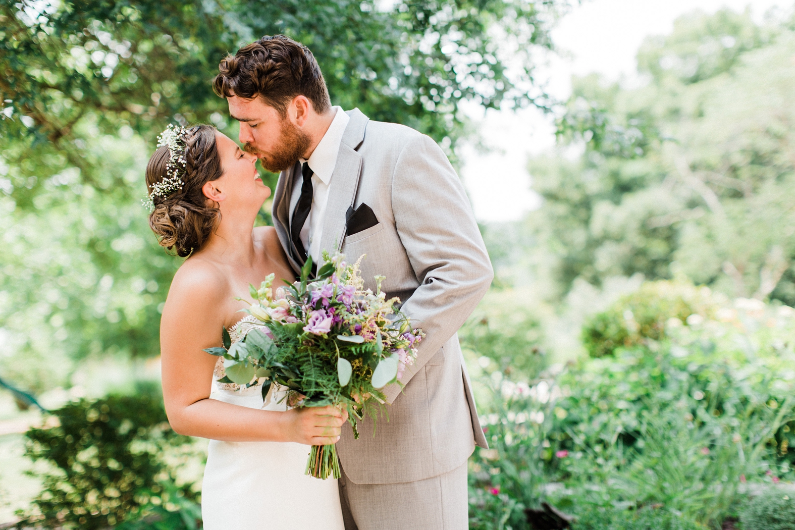 Groom in gray suit with black tie kissing bride on nose; wedding portrait; bride flower crown and purple and succulent bouquet