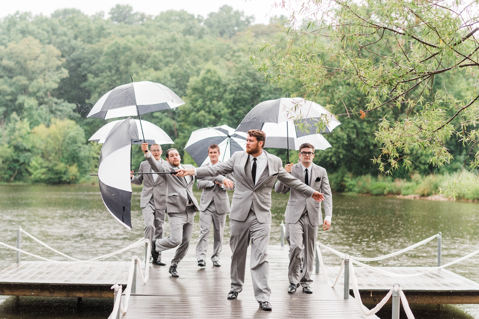 Groom and groomsmen in gray suits with black ties on dock in rain with black and white umbrellas. Little Piney Lodge wedding.