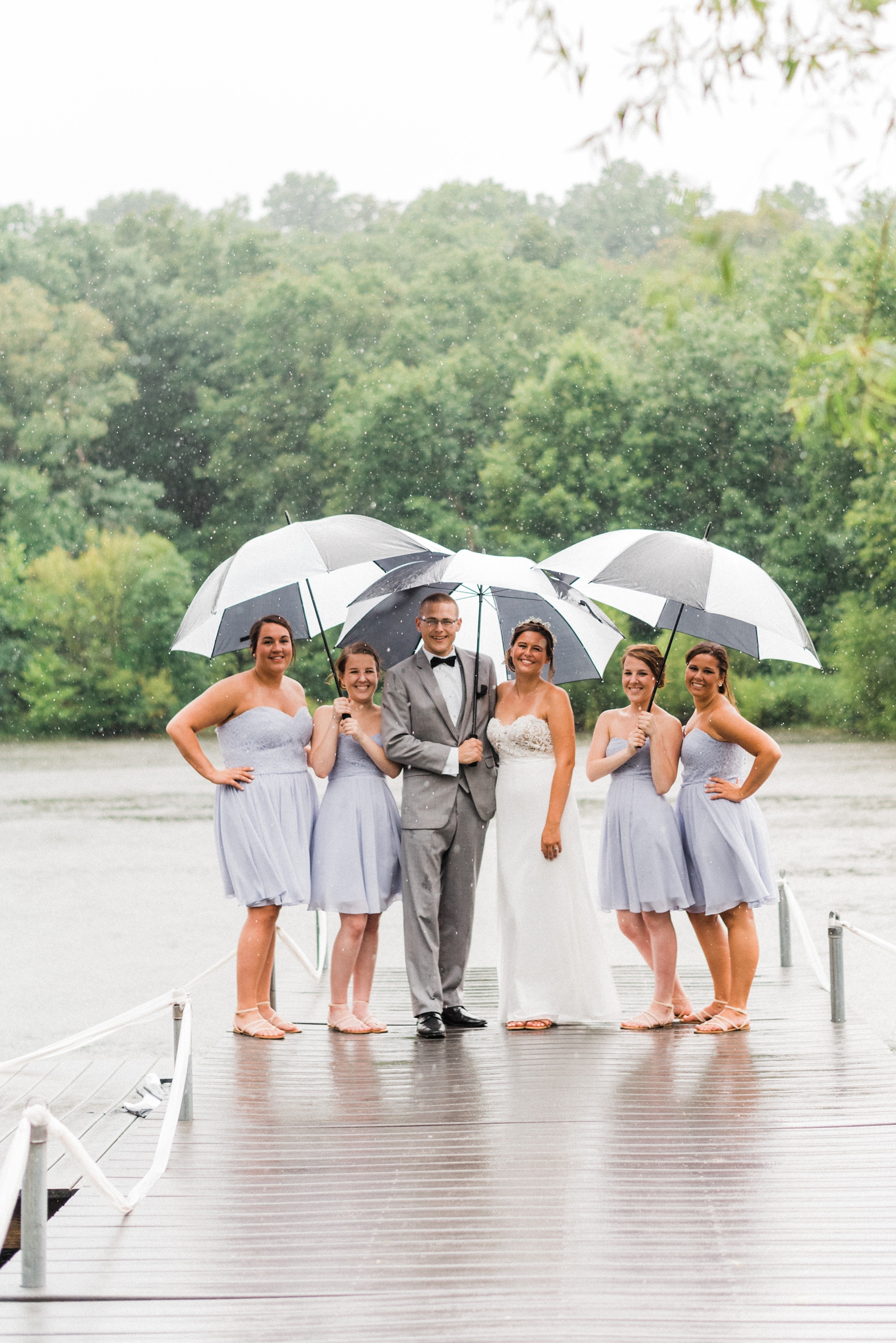 Bride and bridesmaids in rain on dock with black and white umbrellas. Groomsman in gray suit with black tie.