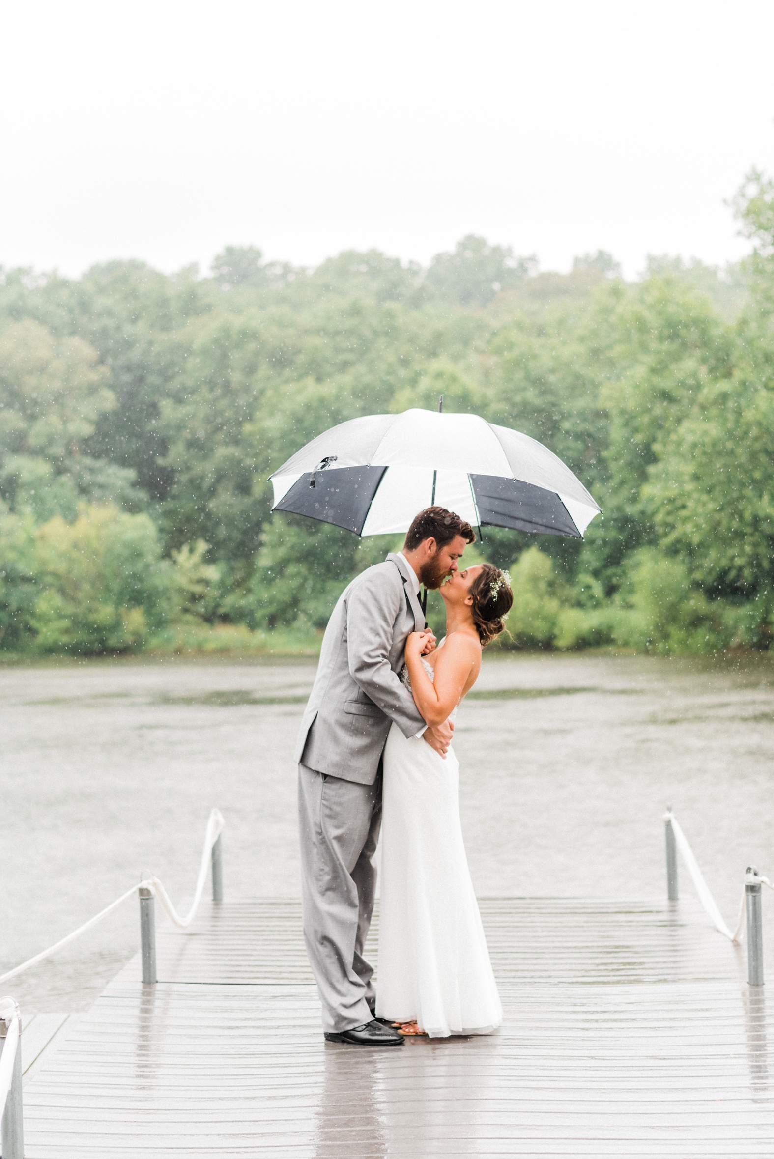 Bride and groom kissing on dock in rain with black and white umbrella. Groom in gray suit with black tie.