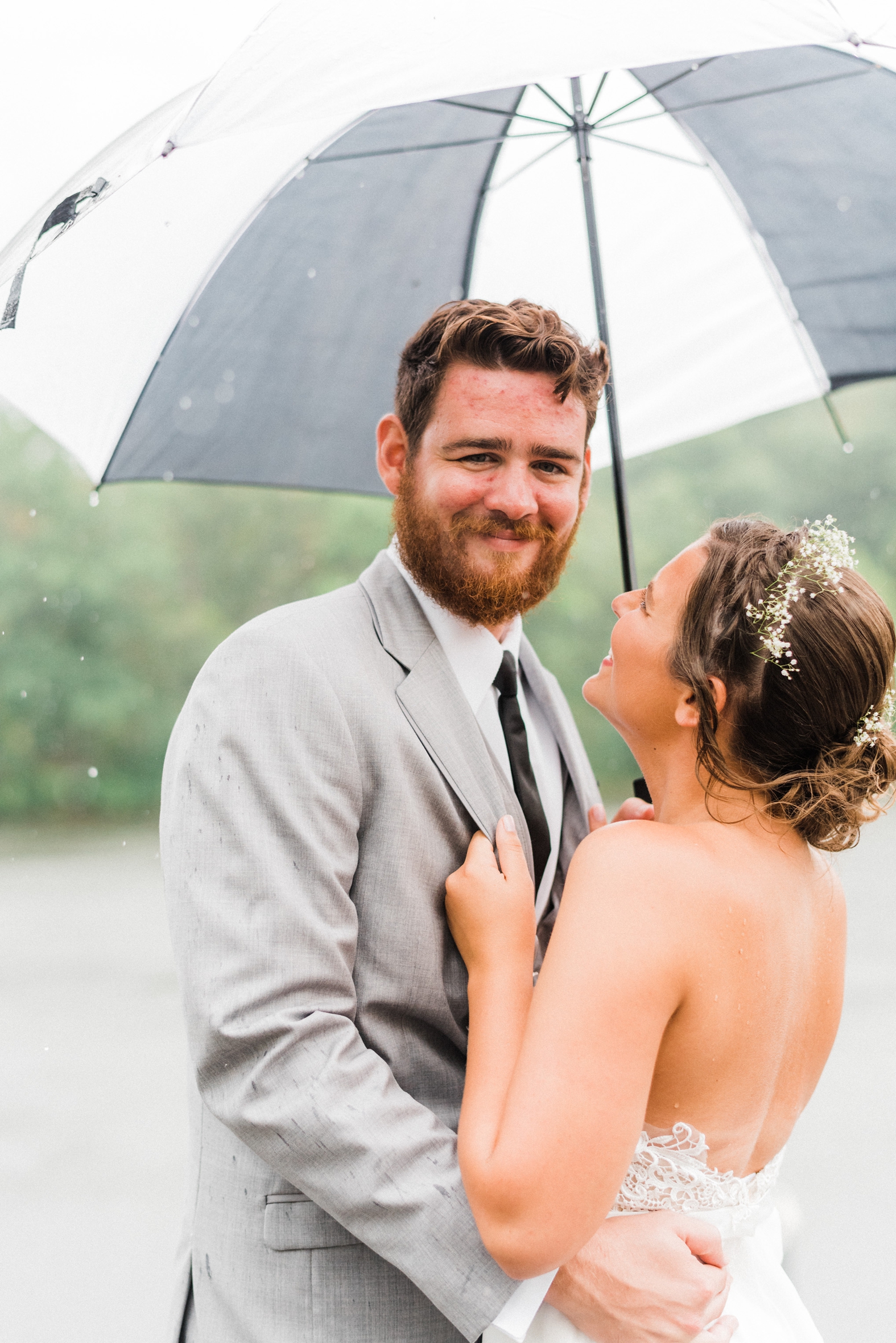 Bride and groom in rain with black and white umbrella. Groom in gray suit with black tie. Bride flower crown with baby's breath.