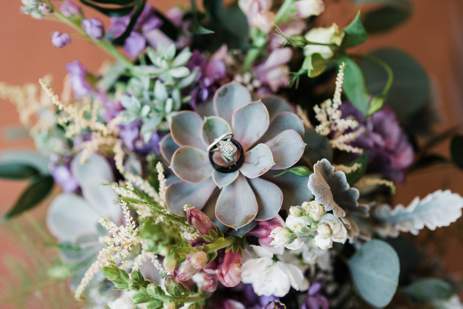 Engagement ring and wedding bands resting on succulent and purple bridal bouquet; wedding details.
