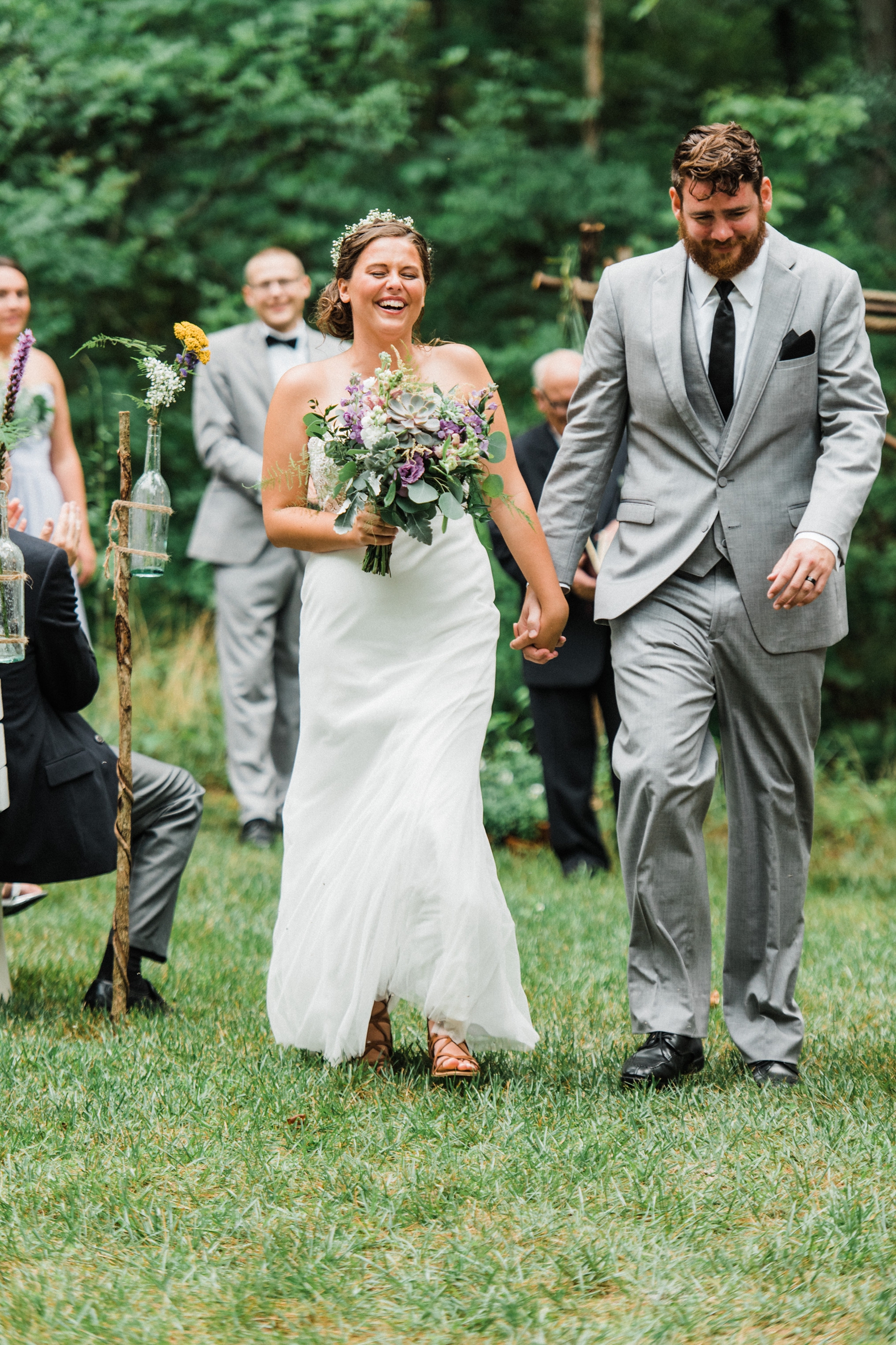 Bride and groom laughing at outdoor wedding ceremony; groom in gray suit with black tie; bride with purple, succulent, and eucalyptus bouquet.