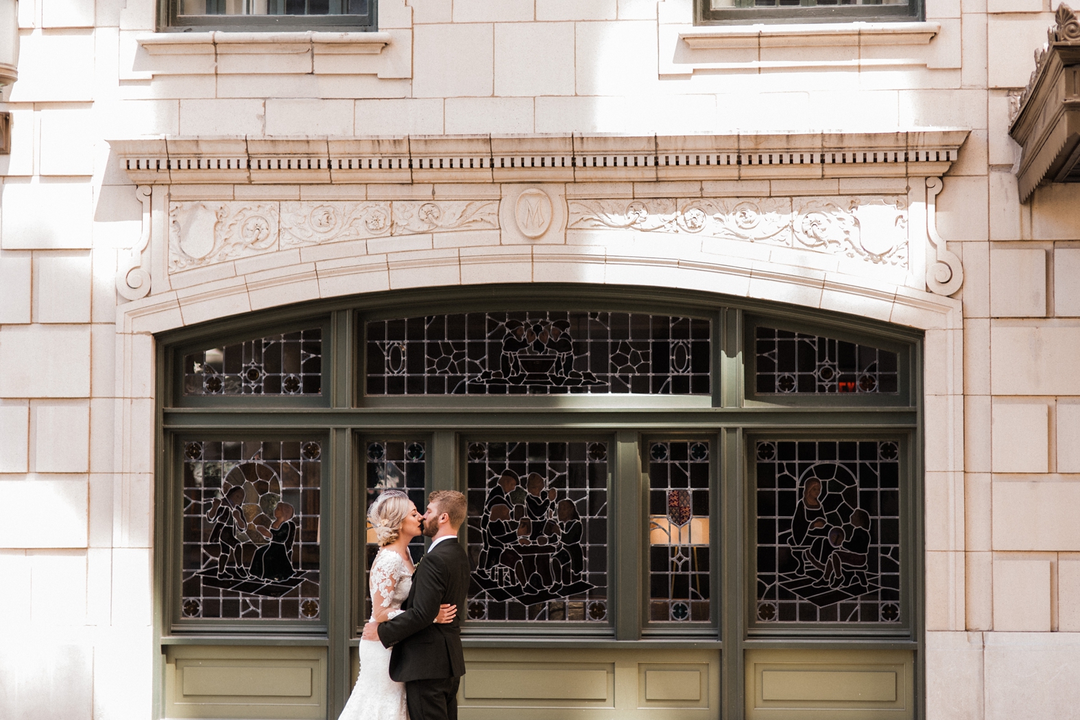 Bride in lace long sleeve wedding dress and birdcage veil; groom in olive green suit kissing in front of stained glass window