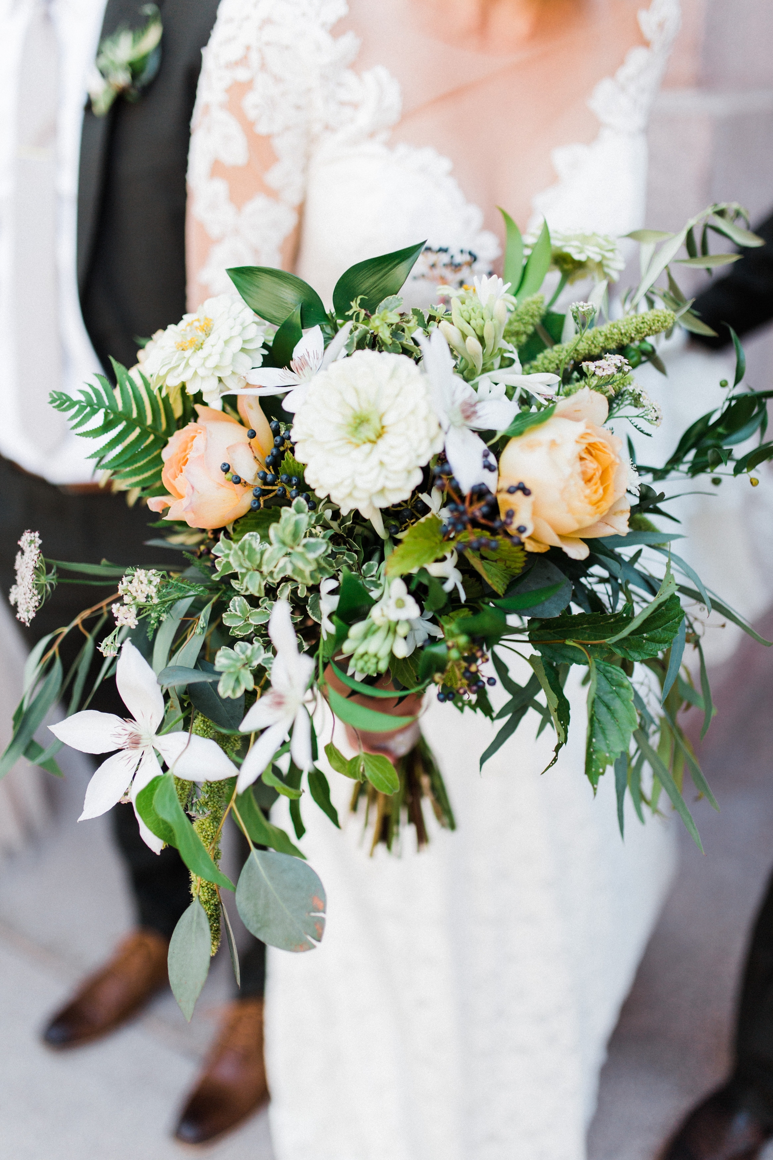 Bridal bouquet with white, ivory, peach roses, daisies, greenery, eucalyptus. Held by bride in lace wedding dress.