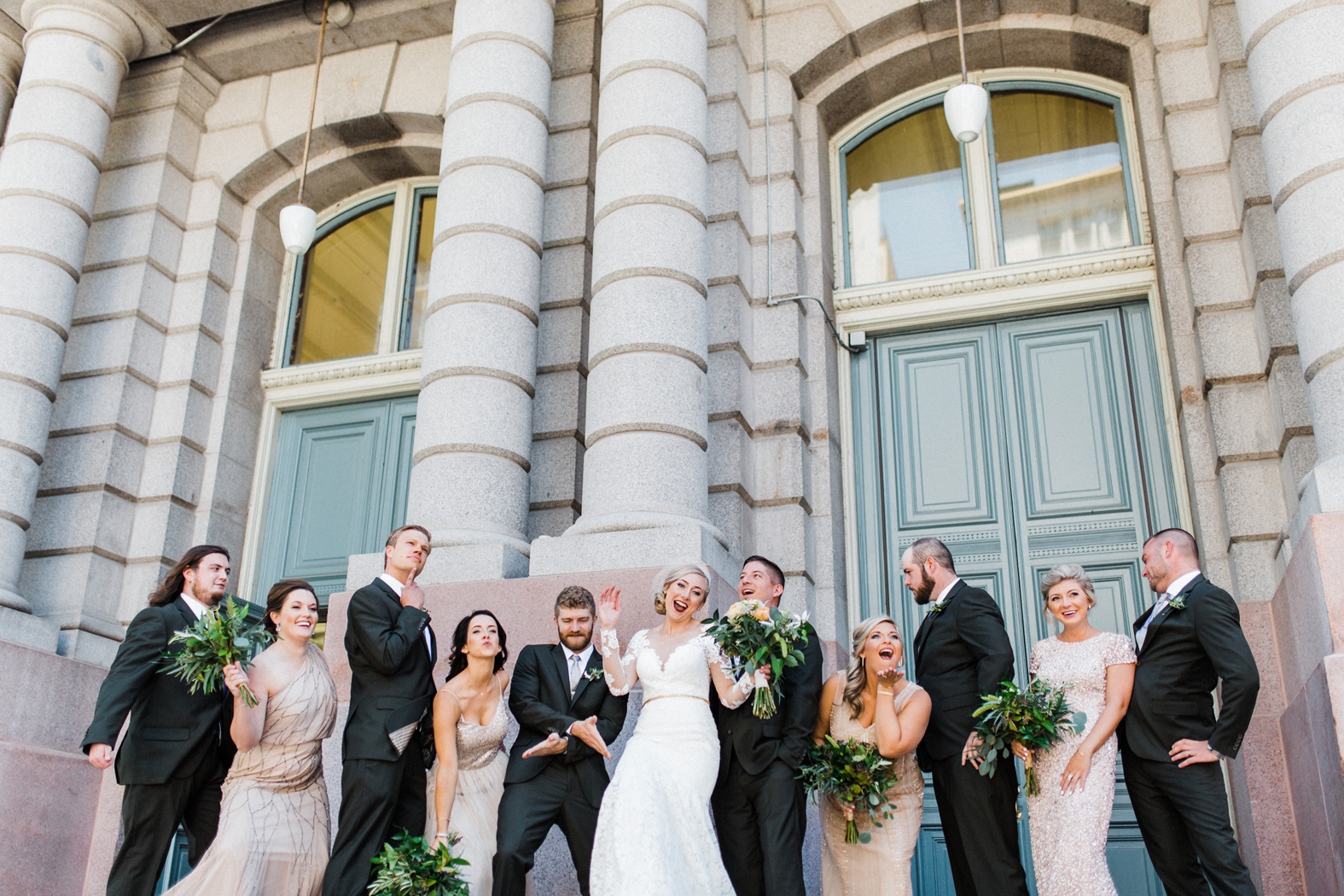 Wedding party silly photo. Groomsmen in olive green suits with light gray ties and brown shoes. Bridesmaids in peach, blush, neutral sequined and beaded bridesmaid dresses. Bride in lace long sleeve wedding gown with birdcage veil. Greenery and peach bouquets.