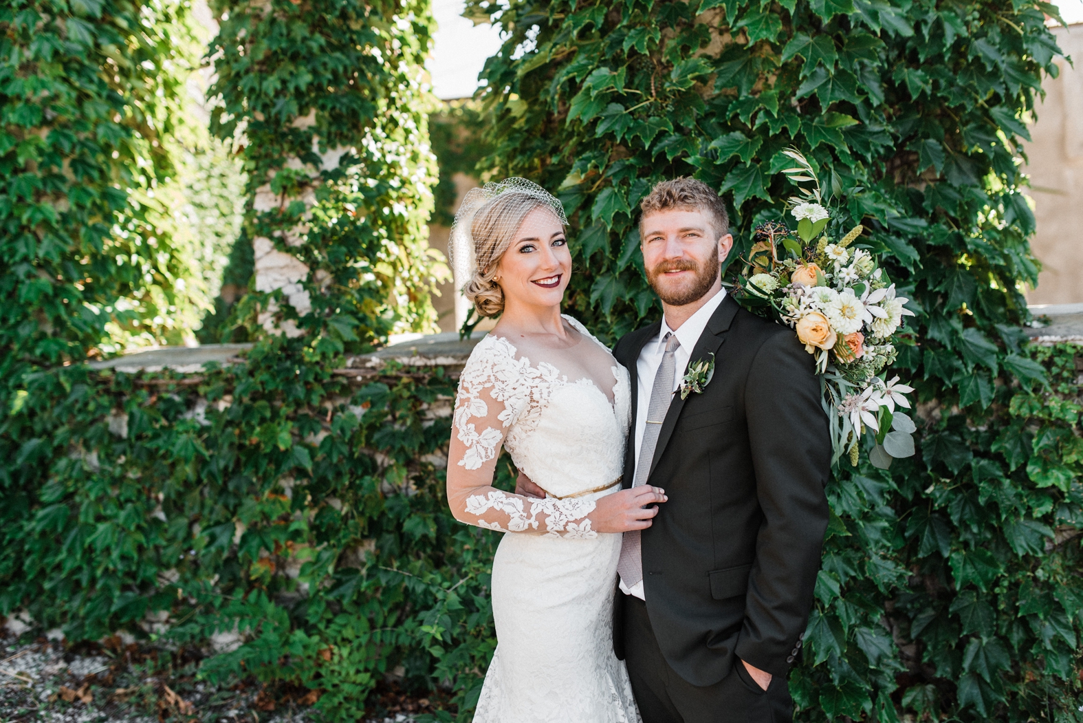 Bride in lace overlay dress with long sleeves, gold belt, birdcage veil. Groom in olive green suit with silver tie, greenery boutonniere. Bridal bouquet with peach ivory and greenery.