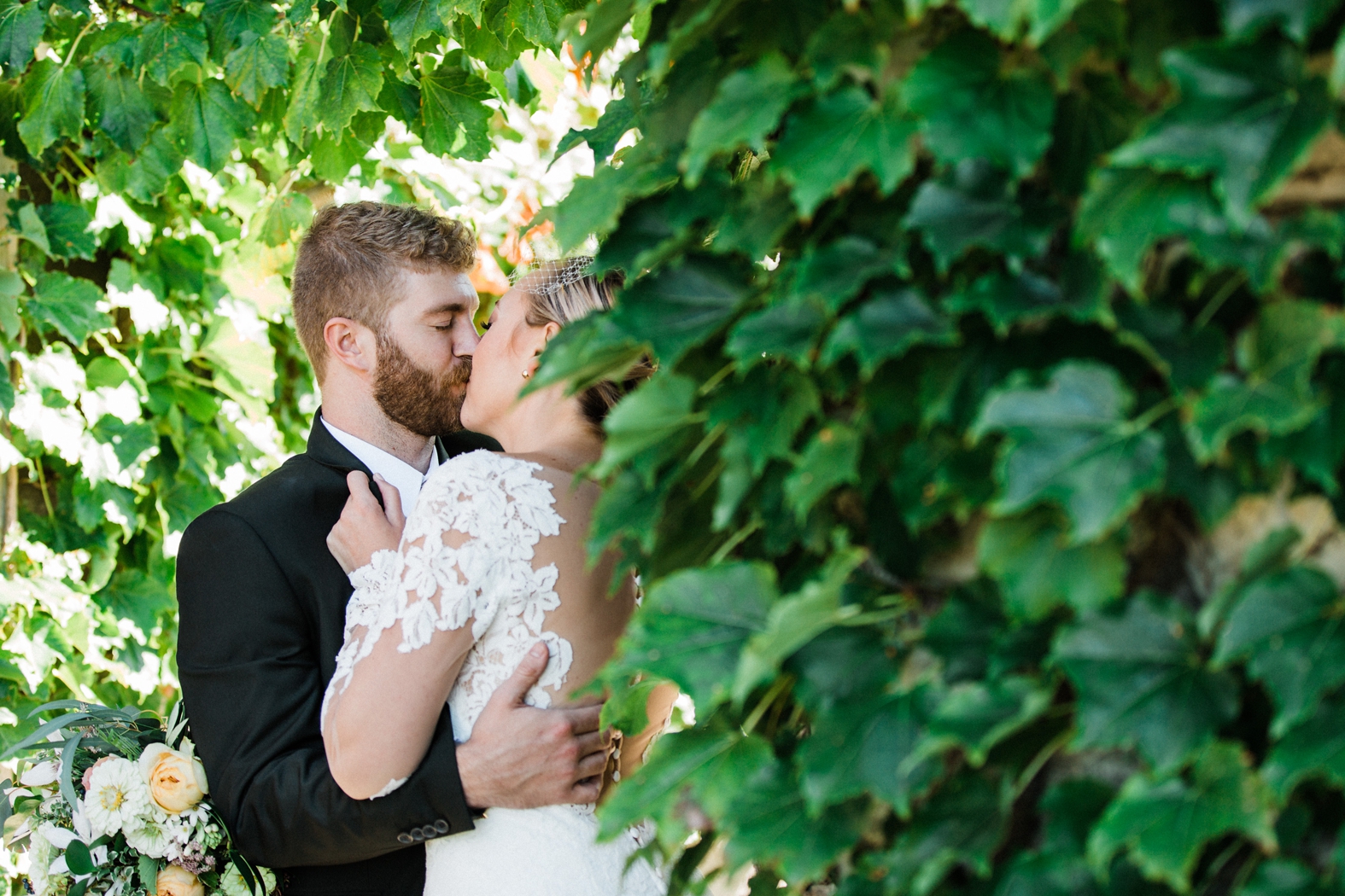 Bride and groom kissing in ivy covered archway. Bride in lace long sleeve backless dress, groom in olive green suit. Peach and ivory bridal bouquet.