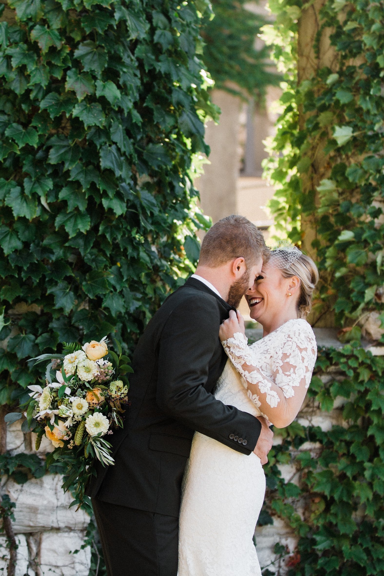 Bride and groom kissing in ivy covered archway. Bride in lace long sleeve backless dress and birdcage veil, groom in olive green suit. Peach and ivory bridal bouquet.