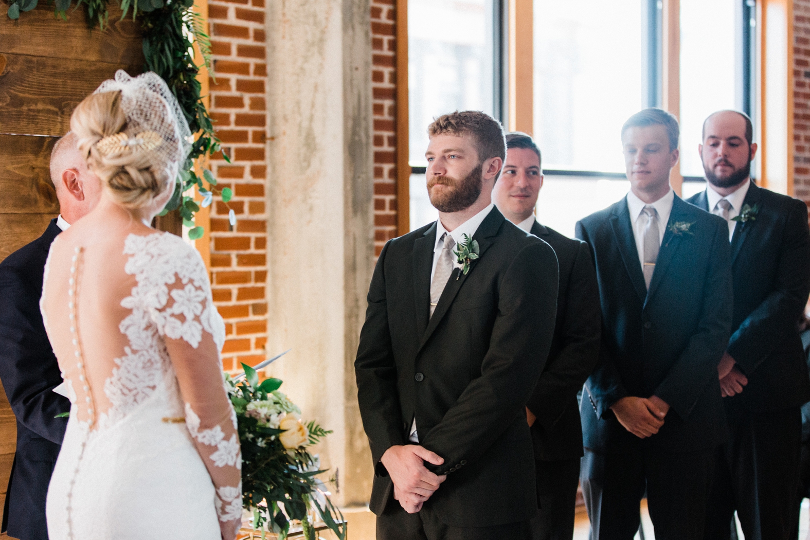Wedding ceremony at Windows on Washington with wood backdrop with greenery. Bride in lace long sleeve wedding dress with buttons up back, birdcage veil, gold hairpiece. Groom in olive green suit with silver tie.