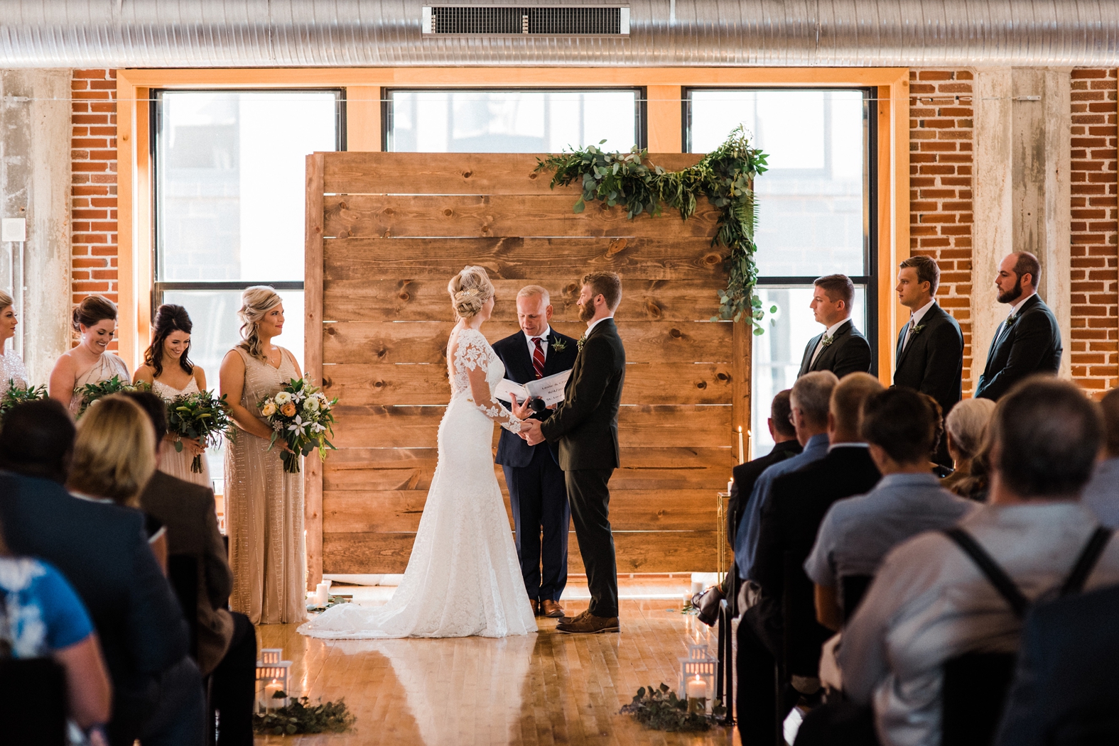 Wedding Ceremony at Windows on Washington with wood backdrop and greenery. Bride in long sleeve lace gown; groom in olive green suit.