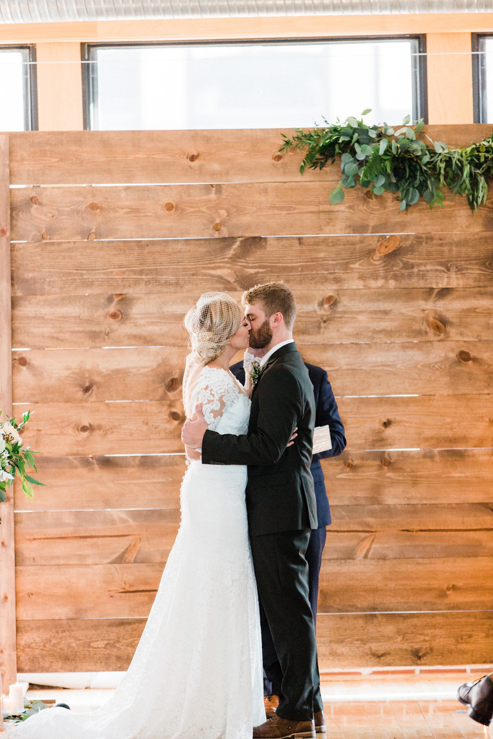Bride and groom kiss at wedding ceremony at Windows on Washington. Wood backdrop with greenery. Bride in lace long sleeve dress with birdcage veil; groom in olive green suit.
