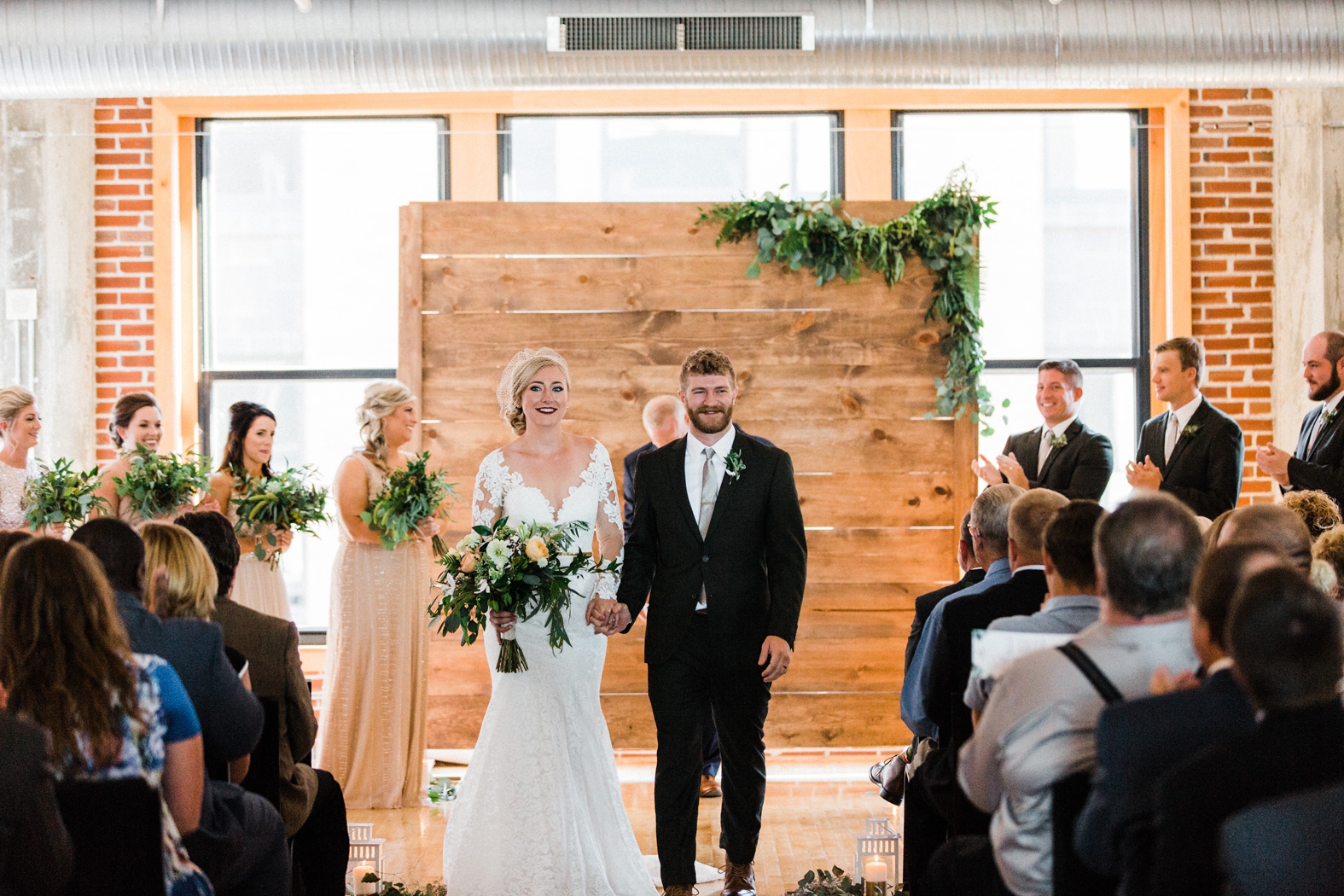 Bride and groom exit wedding ceremony at Windows on Washington. Wood backdrop with greenery. Bride in lace long sleeve dress with birdcage veil; groom in olive green suit.