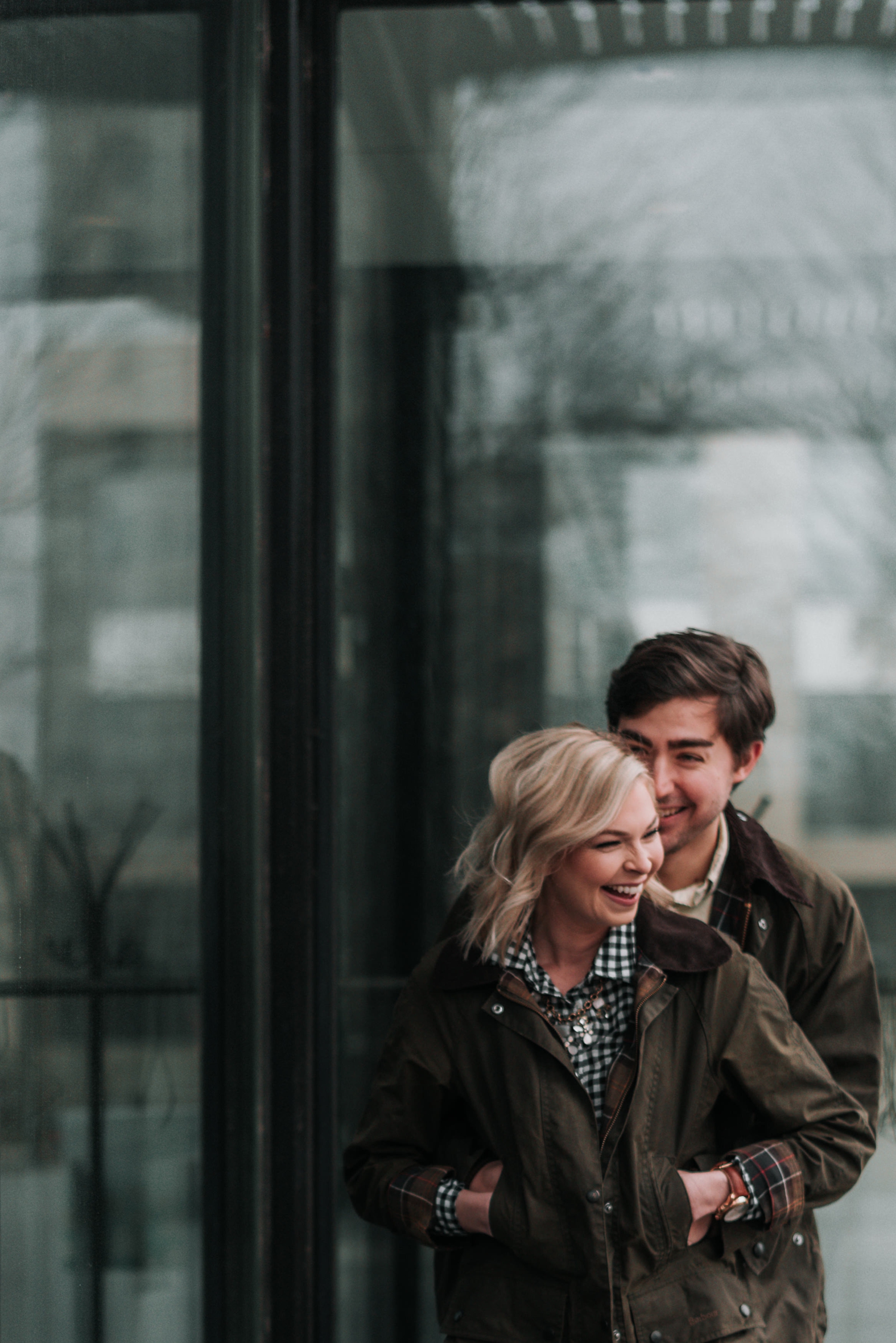 Groom hugs his fiancé from behind in front of glass building at their winter downtown St. Louis engagement photo session.