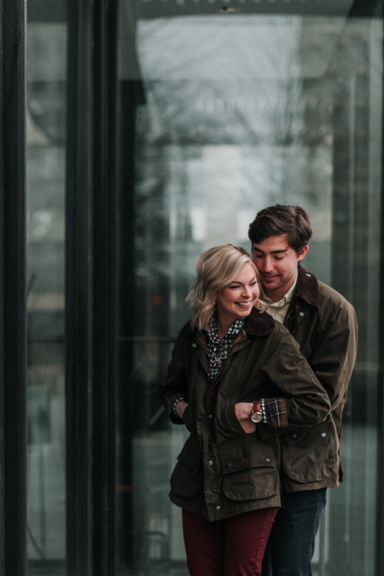 Bride to be laughs as her fiancé hugs her tight from behind at the Peabody Office Building in downtown St. Louis during their winter engagement photo session.