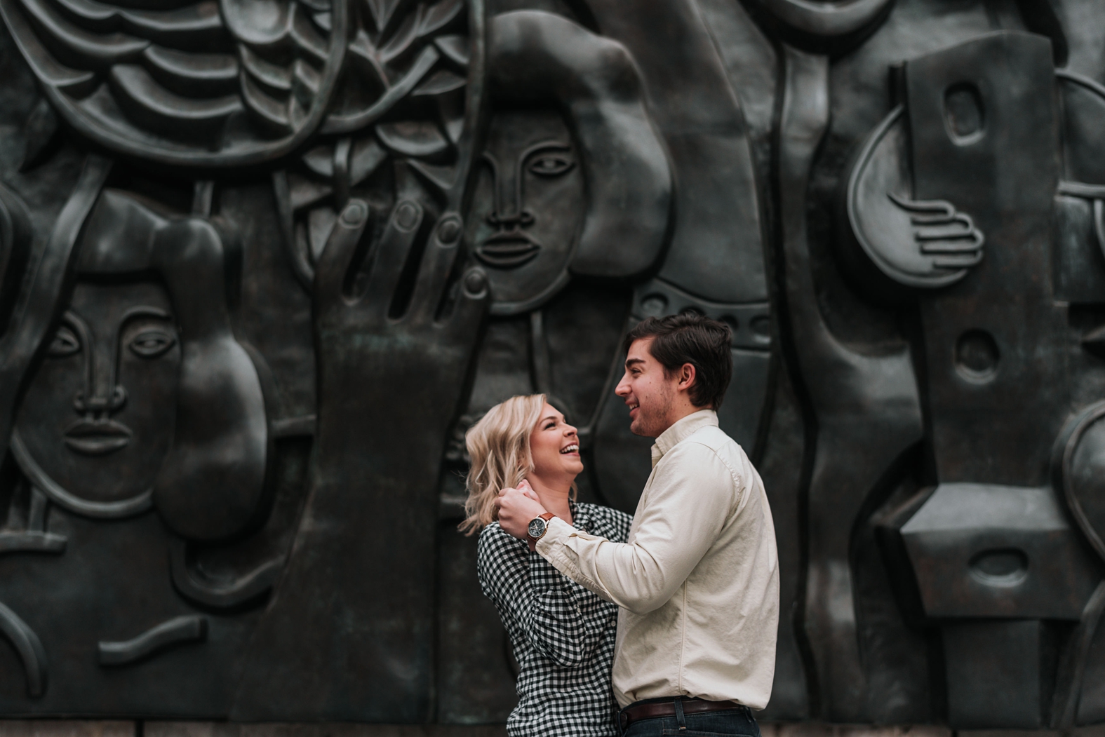 Bride and groom to be dance and laugh in front of black sculpture wall in downtown St. Louis during their winter engagement photo shoot.