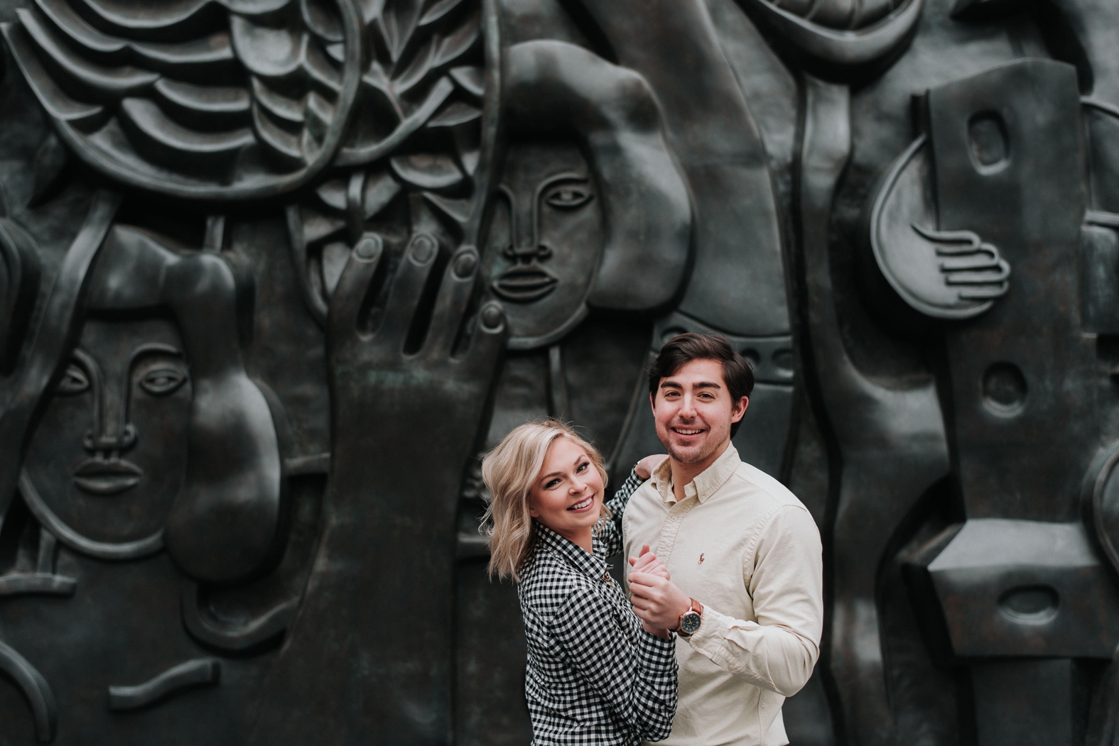 Engaged couple dances and smiles into the camera in front of a black sculpture wall in downtown St. Louis during their winter engagement photos.