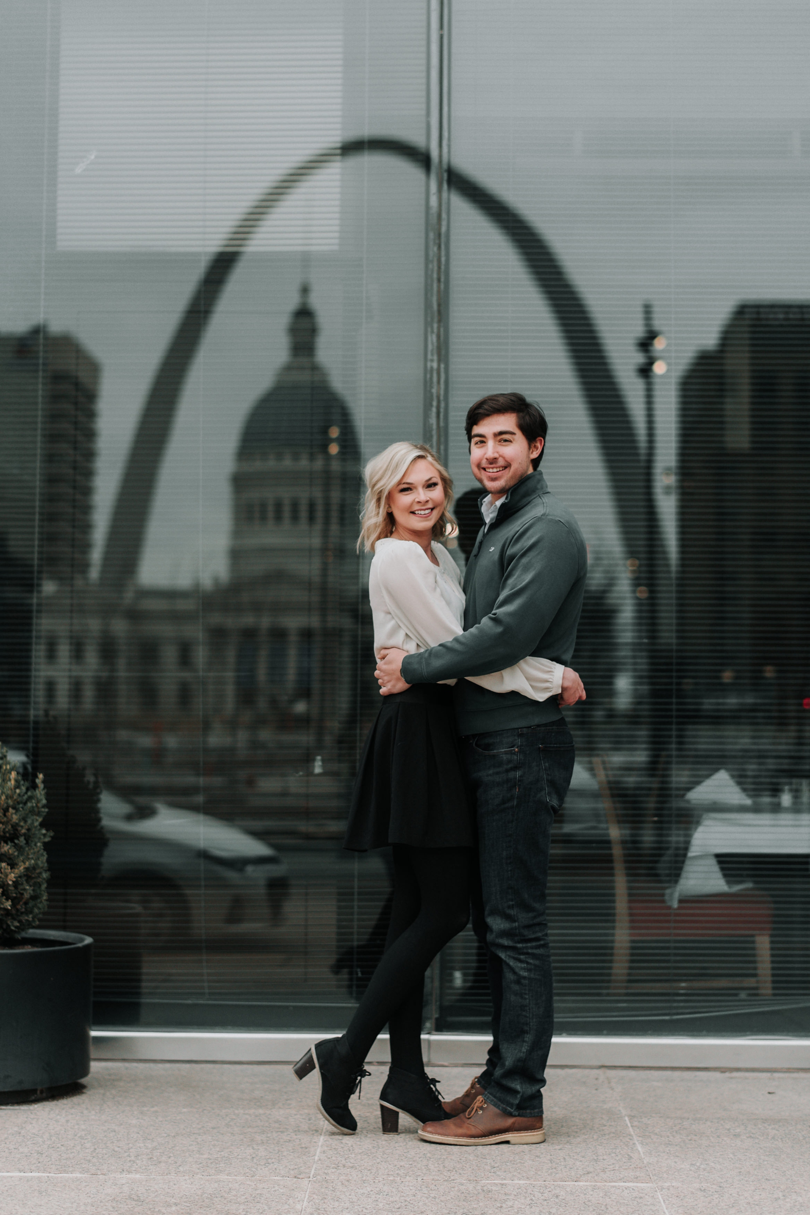 Bride and groom to be hug tightly in front of the reflection of the Gateway Arch in downtown St. Louis during their winter engagement photo shoot.