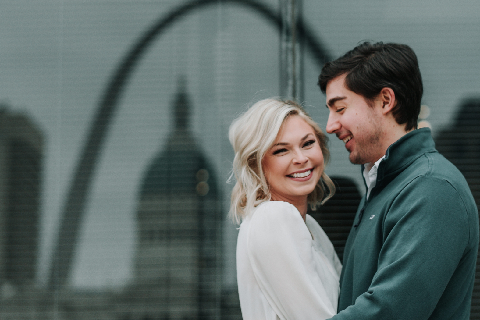 Bride smiles into camera as her groom hugs her close in front of the reflection of the Gateway Arch in a building window in downtown St. Louis during their winter engagement photos.