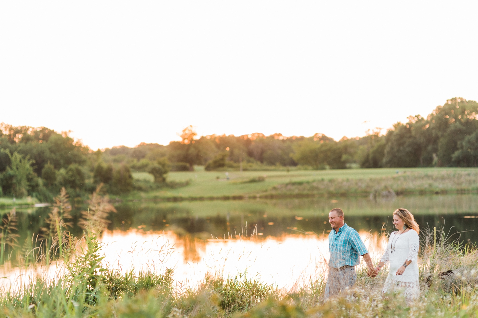 Groom leads bride through a beautiful field by a lake at their engagement photo session in Broemmelsiek Park in St. Charles, MO.